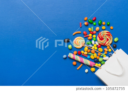 shopping paper gift bag in corner full of assorted traditional candies falling out on colored background with copy space. Happy Holidays sale concept 105872735