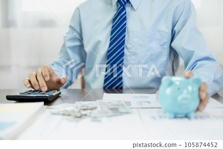 Man calculating her expenses on a notebook putting coin into piggy bank. Saving money with coins Step into a business that is growing to be successful and save for retirement concepts Man calculating her expenses on a notebook putting coin into piggy bank. Saving money with coins Step into a business that is growing to be successful and save for retirement concepts 105874452