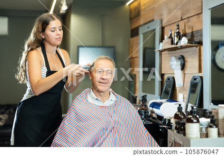 Young woman barber doing haircut with clippers to elderly man in barbershop Young woman barber doing haircut with clippers to elderly man in barbershop 105876047