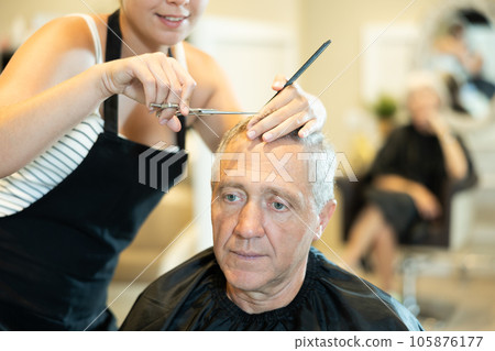 Cropped young girl hairdresser performs haircut for elderly man using scissors Cropped young girl hairdresser performs haircut for elderly man using scissors 105876177