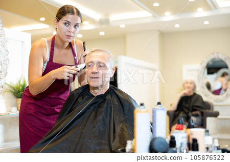Young woman hairdresser performs haircut for elderly man using hair clipper Young woman hairdresser performs haircut for elderly man using hair clipper 105876522