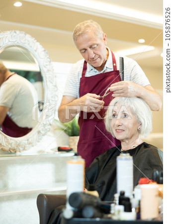 Experienced senior man master makes performs haircut for mature woman with scissors Experienced senior man master makes performs haircut for mature woman with scissors 105876526