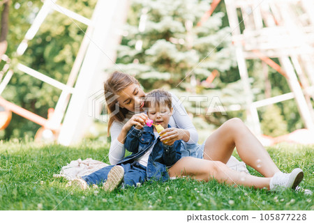 Happy and cheerful mom and son are blowing soap bubbles in a summer park, sitting on the grass 105877228
