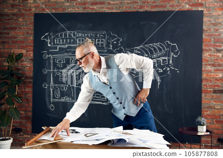 Bearded senior man, architect sitting at table and working with buildings design, sketches. Making measurements, checking 105877808