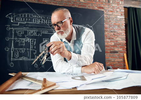 Bearded senior man, architect sitting at table and working with buildings design, sketches. Making measurements 105877809