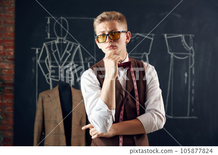 Handsome, stylish young man, fashion designer in classical clothes standing against blackboard with clothing sketches Handsome, stylish young man, fashion designer in classical clothes standing against blackboard with clothing sketches 105877824