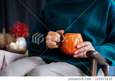 Close up pumpkin shaped cup of hot coffee or tea drink with going steam in female hands with fall mood decor for hygge home on background. Cozy autumn beverage for Thanksgiving or Halloween holidays. 105879216