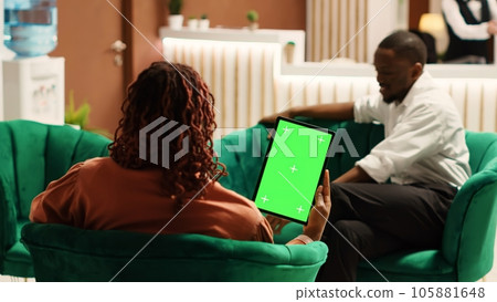 Happy guests in hotel lounge holding chroma key green screen mock up tablet while enjoying cup of coffee. African american couple waiting to be checked in, sitting on cozy sofa Happy guests in hotel lounge holding chroma key green screen mock up tablet while enjoying cup of coffee. African american couple waiting to be checked in, sitting on cozy sofa 105881648