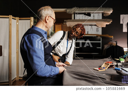 Helpful old master dressmaker guiding happy apprentice with cutting textile material for upcoming bespoken sartorial collection. Couturiers in tailoring studio working on making stylish fashion attire 105881669