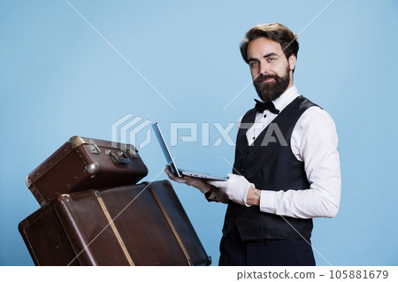 Doorman with laptop in hand showing expression of hotel industry, showcases proficiency in utilizing technology to offer assistance. Bellhop in suit making reservations or provides information. 105881679