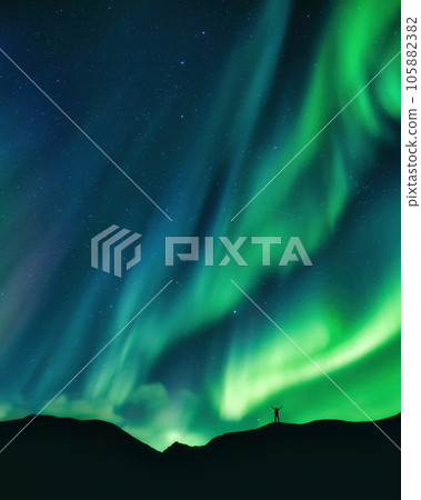 Aurora borealis glowing over hiker standing on mountain in arctic circle Aurora borealis glowing over hiker standing on mountain in arctic circle 105882382