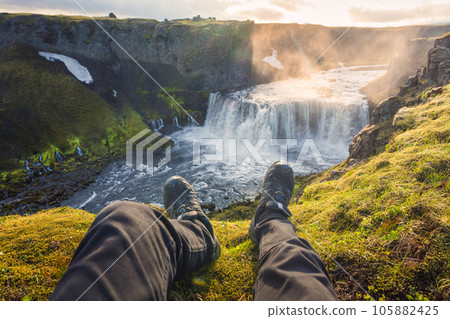 Legs of man sitting on edge of mountain with Axlafoss waterfall flowing in canyon at Icelandic Highlands 105882425