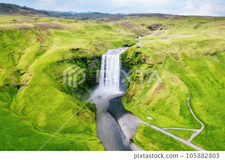 Skogafoss waterfall flowing from cliff surrounded by lush landscape in summer 105882803