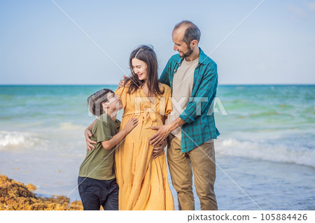 A loving family enjoying tropical beach - a radiant pregnant woman after 40, embraced by her husband, and accompanied by their adult teenage son, savoring precious moments together amidst nature's 105884426