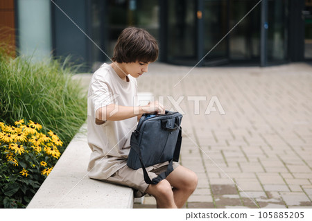 Boy open laptop bag and takes out his laptop. Boy sits on bench outdoors 105885205