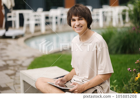 Teenage boy playing games on digital tablet and sitting on a bench under a house in a residential area Teenage boy playing games on digital tablet and sitting on a bench under a house in a residential area 105885208