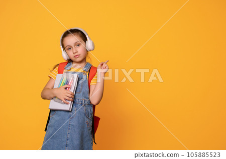 Smart first grade student girl in headphones and casual denim overalls, points aside a copy advertising space for text Smart first grade student girl in headphones and casual denim overalls, points aside a copy advertising space for text 105885523