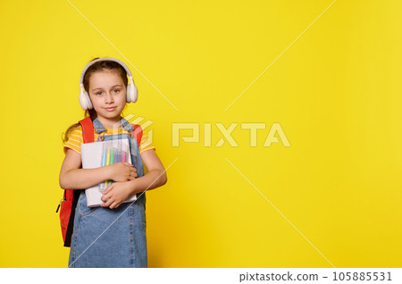 Pretty little girl wearing wireless headphones, carrying a backpack, looking at camera, isolated orange background 105885531