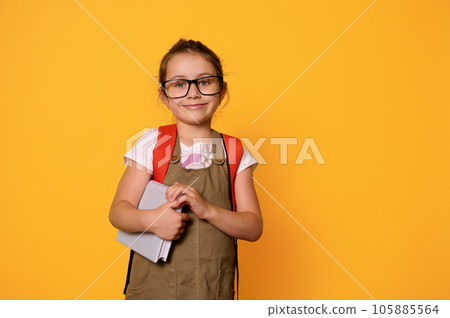 Studio portrait of Caucasian happy smiling elementary age school girl with backpack, holding book and looking at camera 105885564