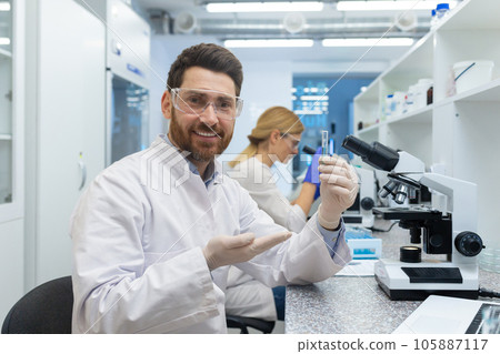 Portrait of a young smiling man working in a laboratory under a microscope, holding and showing a flask with a substance to the camera. A female colleague works next to the table. Portrait of a young smiling man working in a laboratory under a microscope, holding and showing a flask with a substance to the camera. A female colleague works next to the table. 105887117