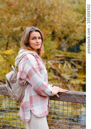 Portrait Of Cute Young Woman In Casual Wear In Autumn, Standing On Bridge Against Background Of An Autumn Park And River. Pretty Female Walking In Park In Golden Fall. Copy Space. Smiling Girl In The 105888302