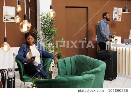 African American woman traveler sitting in hotel lobby with smartphone chatting online with friends while waiting for hotel check-in. People tourists with luggage wait at reception area 105890286