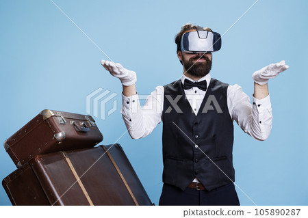 Modern doorkeeper trying on vr headset against blue background, using virtual reality technology in studio. Young man with bellhop occupation standing next to luggage, tourism concept. 105890287