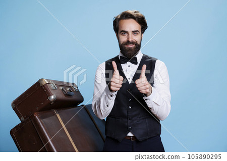 Smiling bellboy does thumbs up sign while he is wearing formal hotel attire in studio, expressing success and agreement. Young man dressed as doorman shows like symbol, tourism industry. Smiling bellboy does thumbs up sign while he is wearing formal hotel attire in studio, expressing success and agreement. Young man dressed as doorman shows like symbol, tourism industry. 105890295