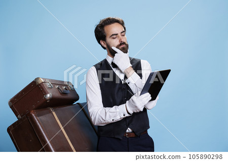 Male porter works with tablet, being in charge of luggage for safekeeping. Doorkeeper conveys his commitment in creating a welcoming atmosphere, symbolizing his role at luxurious hotel. 105890298