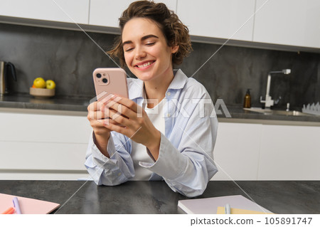 Technology and lifestyle. Young woman sits at home, uses smartphone in her kitchen and smiles 105891747