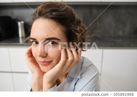Close up portrait of young woman, 25 years old, sitting in her kitchen alone, express candid happiness, smiles, leans head on hands Close up portrait of young woman, 25 years old, sitting in her kitchen alone, express candid happiness, smiles, leans head on hands 105891988