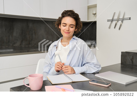 Portrait of young woman working from home, writing down information in notebook, taking notes, sitting in kitchen and studying, student doing homework 105892022