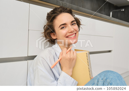 Portrait of modern carefree girl, sits on floor with notebook, smiles at camera, writes in her diary, holds journal in hands 105892024