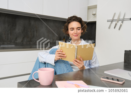 Young woman sitting in kitchen, reading her notes in notebook. Smiling girl at home, doing homework, studying Young woman sitting in kitchen, reading her notes in notebook. Smiling girl at home, doing homework, studying 105892081