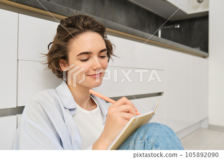 Close up portrait of young woman writing in her journal, feeling creative, sitting with planner, notebook and pen on floor, studying, doing her homework at home 105892090