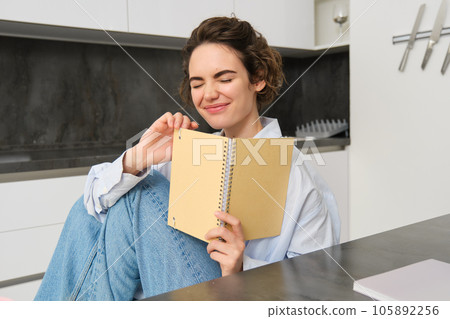 Portrait of carefree brunette woman, laughing and smiling while reading funny text in notebook, reading journal Portrait of carefree brunette woman, laughing and smiling while reading funny text in notebook, reading journal 105892256