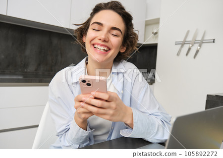 Image of young woman using her smartphone at home. Girl sits with mobile phone in kitchen and smiles 105892272