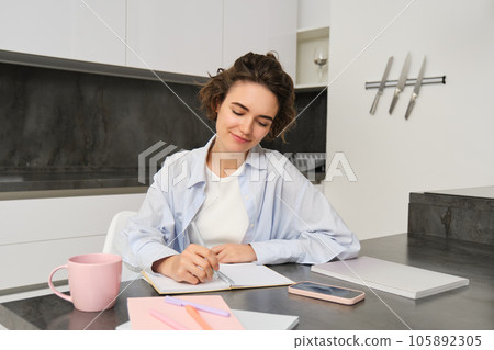 Portrait of young woman working from home, writing down information in notebook, taking notes, sitting in kitchen and studying, student doing homework 105892305