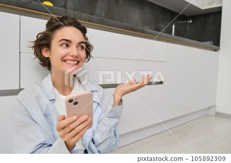 Portrait of happy young woman, shows her brand new kitchen, sits on floor, points at furniture and smiles, video chats on smartphone Portrait of happy young woman, shows her brand new kitchen, sits on floor, points at furniture and smiles, video chats on smartphone 105892309