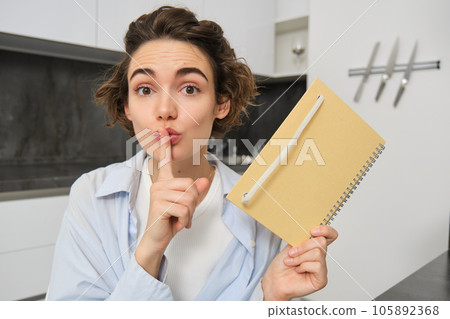 Young woman holds her diary, shows secret, hush gesture, presses finger to lips, puts her thoughts in writing Young woman holds her diary, shows secret, hush gesture, presses finger to lips, puts her thoughts in writing 105892368