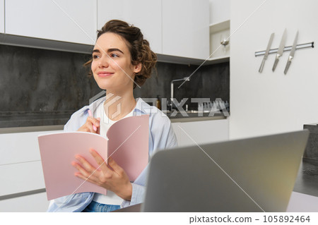 Young woman checking her schedule while working on remote from home, using laptop, looking at her daily planner, making notes, writing down information in notebook 105892464