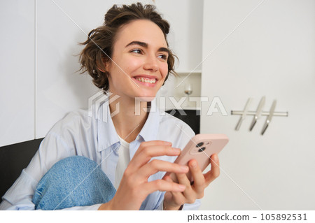 Portrait of smiling beautiful woman, sitting in kitchen with smartphone, looking happy at camera, spending time at home, using mobile phone app 105892531