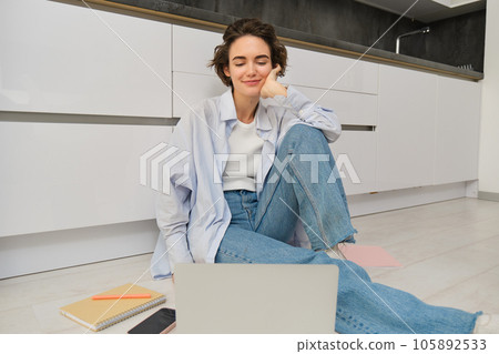 Portrait of smiling young woman watching video on laptop, sitting on kitchen floor and looking at computer screen, connects to online chat 105892533