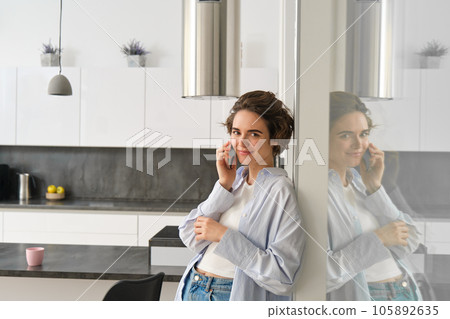 Beautiful woman talking on mobile phone, answers phone call and smiles, stands at home, leans to wall and chats Beautiful woman talking on mobile phone, answers phone call and smiles, stands at home, leans to wall and chats 105892635