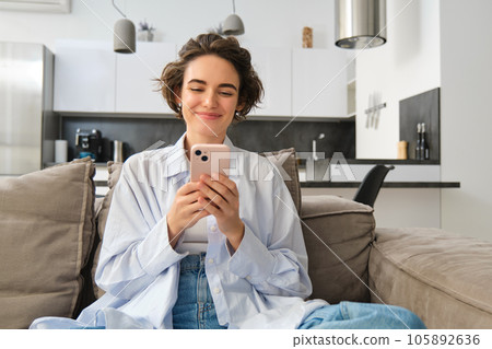 Portrait of smiling woman chats on smartphone app, sits at home on sofa and uses mobile phone application Portrait of smiling woman chats on smartphone app, sits at home on sofa and uses mobile phone application 105892636
