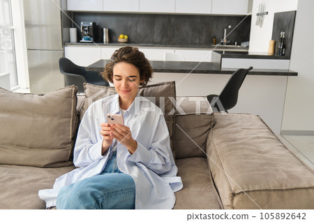 Portrait of brunette woman sits on sofa with smartphone, chats on phone, looks at her mobile app, checks messages on telephone, does online shopping from home Portrait of brunette woman sits on sofa with smartphone, chats on phone, looks at her mobile app, checks messages on telephone, does online shopping from home 105892642