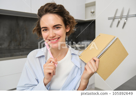 Smiling brunette womnan, holding her diary, writing down in planner, working from home on laptop, studying online and taking notes in notebook Smiling brunette womnan, holding her diary, writing down in planner, working from home on laptop, studying online and taking notes in notebook 105892649
