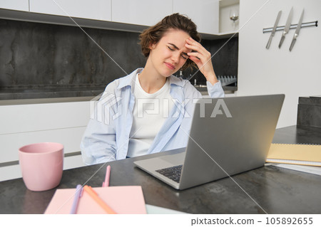 Woman has headache while works from home. Girl has painful migraine, sits near laptop, grimaces from discomfort Woman has headache while works from home. Girl has painful migraine, sits near laptop, grimaces from discomfort 105892655