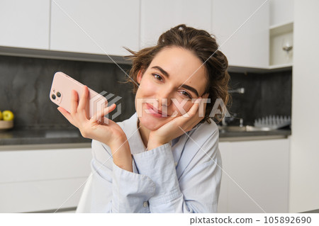 Candid smiling woman, holding smartphone in her kitchen, using mobile phone and looking happy 105892690