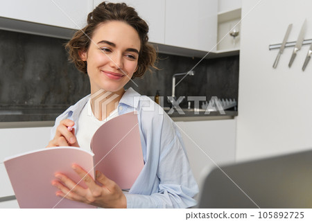 Young smiling woman works from home, writes in her diary, makes notes in journal planner, sits near laptop in kitchen 105892725